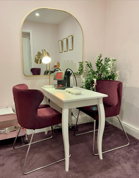 permanent/welded jewellery studio with two purple chais opposite sides of a white desk with a small welding machine and mirror hanging on a pink wall